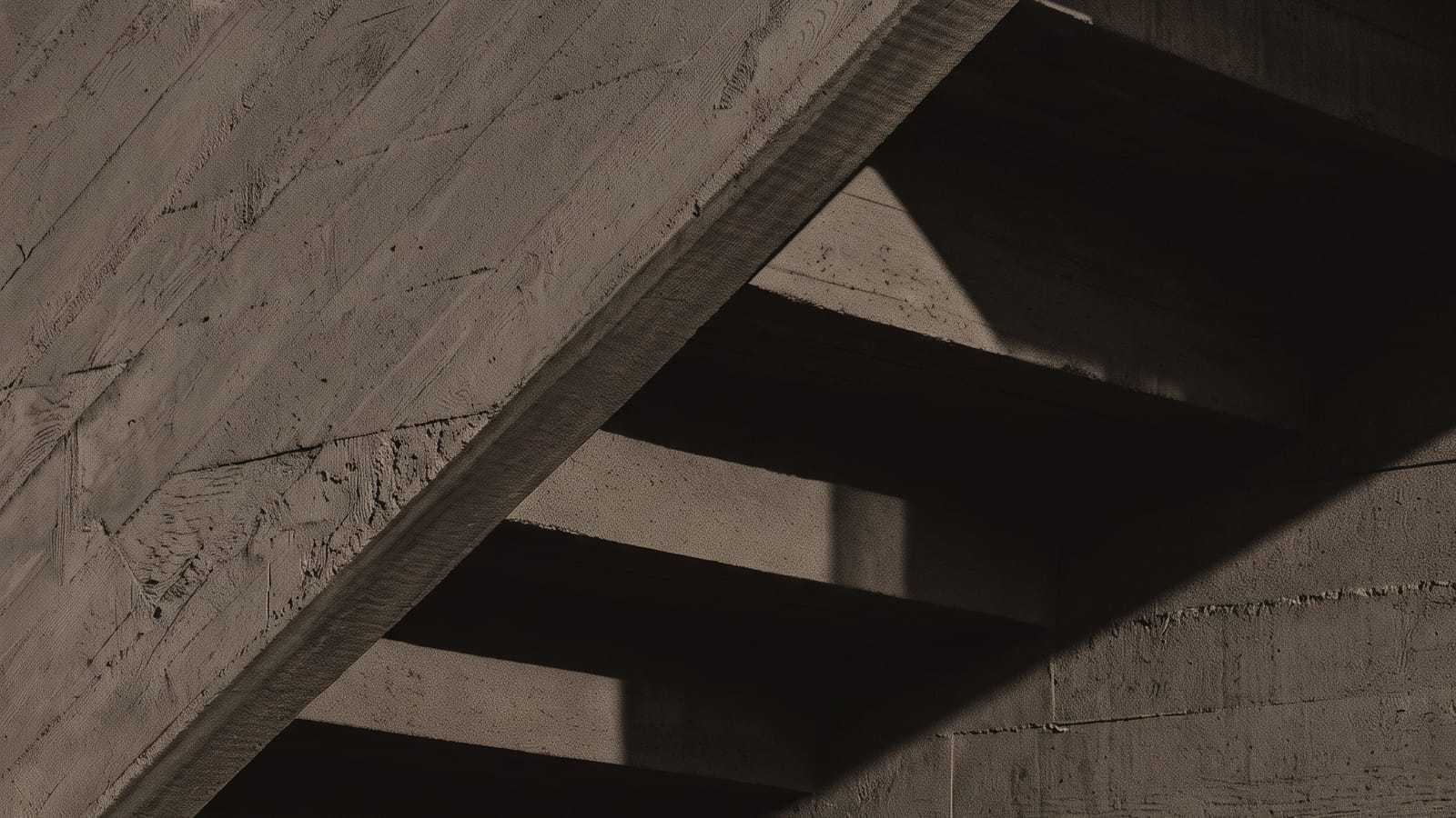 Die Unterseite einer Betontreppe, von der Seite angestrahlt. Die Abdrücke der Brettschalung sind zu sehen, eine harte Schattenkante läuft quer durch das Bild.